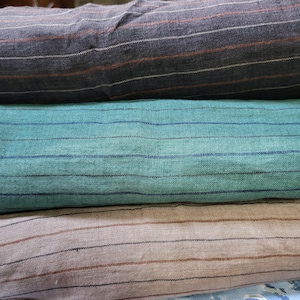 May include: Three folded layers of woven fabric stacked vertically. The top layer is dark charcoal gray with thin white and rust-colored stripes. The middle layer is teal green with thin dark blue stripes. The bottom layer is light beige with thin brown stripes.