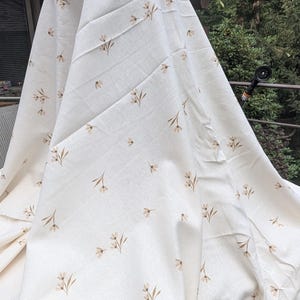 May include: A large piece of off-white fabric featuring a repeating pattern of small, light brown floral sprigs. The textile has a visible woven texture and is draped in soft folds, showcasing the delicate botanical print across the surface.