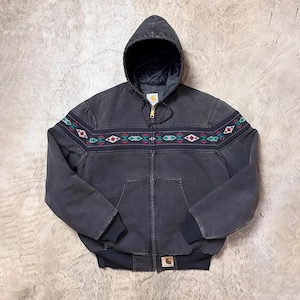 Op de afbeelding: Een donkergrijze jas met capuchon, een ritssluiting over de hele lengte en een decoratief geometrisch patroon in teal, rood en wit over de borst en mouwen. De jas heeft een zwarte geribde zoom en manchetten en een kleine logopatch.