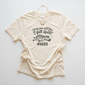 Könnte beinhalten: Cremefarbenes T-Shirt mit dem Text "every now and then I fall apart" über einer Zebra-Illustration und dem Hashtag #hEDS. Eine goldene Halskette liegt auf dem Shirt.
