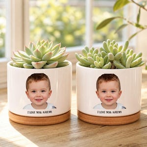 May include: Two white cylindrical ceramic planters with wooden bases, each containing a green succulent. The front of each planter features a color photograph of a young child and the printed text "I LOVE YOU. KAILYN" in black lettering.