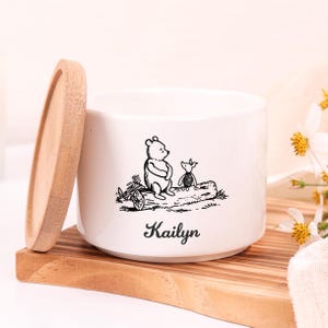 Peut inclure: Un récipient en céramique blanc avec un couvercle en bois, orné d'une illustration noire de Winnie l'ourson et Porcinet. Le nom "Kailyn" est imprimé en dessous. Le récipient est posé sur un plateau en bois.
