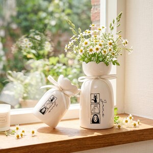 May include: Two white ceramic vases with black illustrations and the word "June" in script. One vase holds a bouquet of white flowers. The other vase is tied with a white ribbon. The vases are on a wooden windowsill.