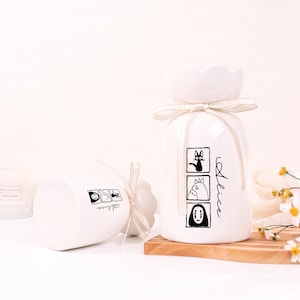 May include: Two white ceramic vases with a flower-shaped top and a ribbon. One vase is upright, the other is on its side. Each vase has a black illustration and the name "Alice" written on it. The upright vase has three illustrations.
