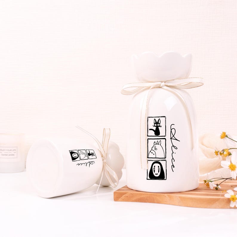 May include: Two white ceramic vases with a flower-shaped top and a ribbon. One vase is upright, the other is on its side. Each vase has a black illustration and the name "Alice" written on it. The upright vase has three illustrations.