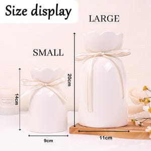 May include: Two white ceramic vases, one small and one large, with a flower-shaped top and tied with a ribbon. The small vase is 14cm tall and 9cm wide, while the large vase is 20cm tall and 11cm wide. The text "Size display" is at the top.