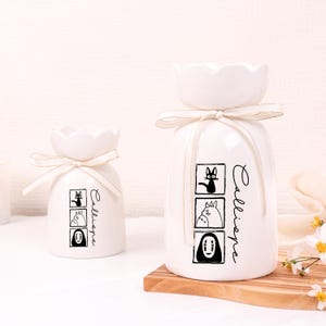 May include: Two white ceramic vases with a flower-shaped top and a cream-colored ribbon. Each vase features black and white cartoon character illustrations and the word "Calliope" in script.