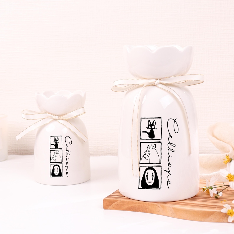 May include: Two white ceramic vases with a flower-shaped top and a cream-colored ribbon. Each vase features black and white cartoon character illustrations and the word "Calliope" in script.