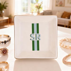 May include: A square white ceramic trinket dish with a gold-toned rim. The center features a vertical design consisting of two dark green stripes flanking a light blue stripe, with the letters SR in light blue and dark green layered over the center.
