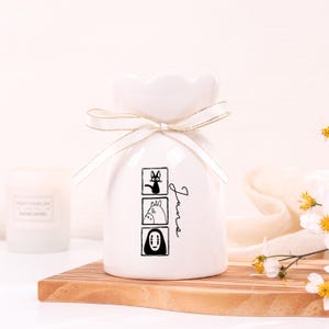 May include: White ceramic vase with a ruffled top, tied with a gold-edged ribbon. The vase features three black and white cartoon character illustrations and the word "June" in script. The vase is on a wooden board.