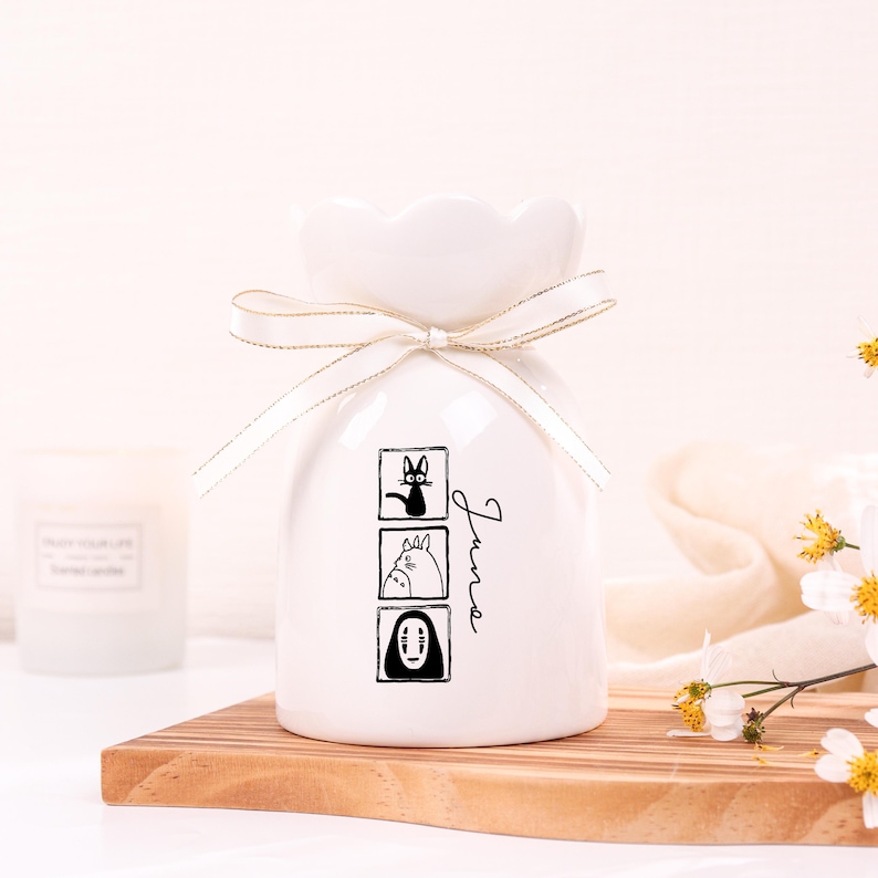 May include: White ceramic vase with a ruffled top, tied with a gold-edged ribbon. The vase features three black and white cartoon character illustrations and the word "June" in script. The vase is on a wooden board.