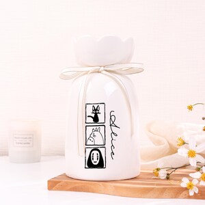 May include: White ceramic vase with a flower-shaped top and a cream-colored ribbon. The vase features three black and white cartoon character illustrations and the name "Alice" in script. The vase is on a wooden surface.