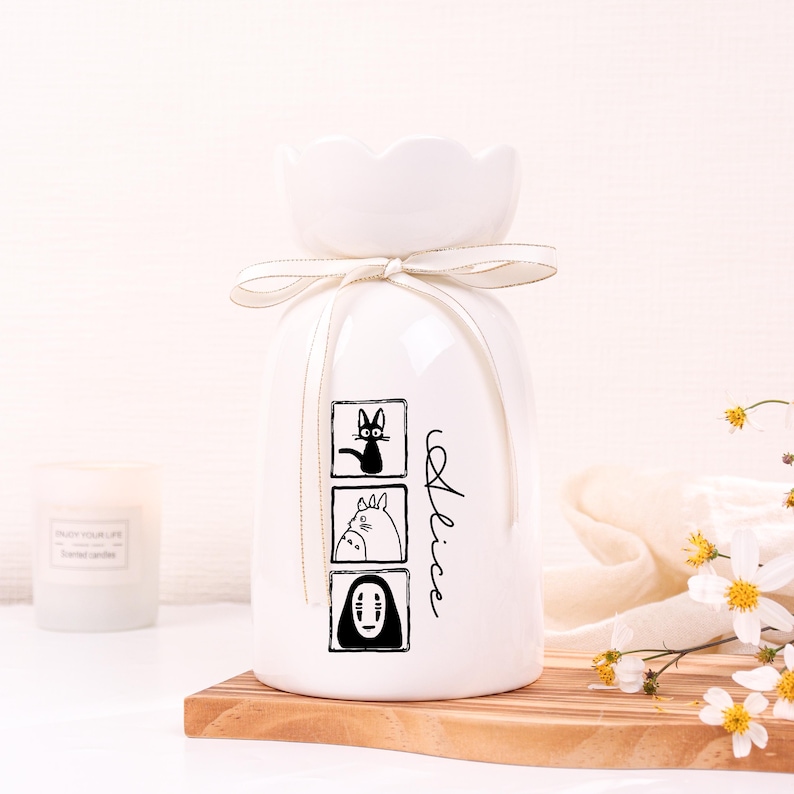 May include: White ceramic vase with a flower-shaped top and a cream-colored ribbon. The vase features three black and white cartoon character illustrations and the name "Alice" in script. The vase is on a wooden surface.