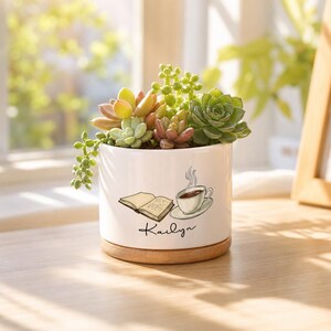 May include: A white ceramic cylindrical planter with a wooden base, containing a variety of green and reddish succulents. The planter features a graphic of an open book and a steaming cup of coffee, with the name Kailyn printed in black script.