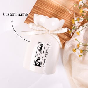 May include: A white ceramic vase with a scalloped top and a cream-colored ribbon. The vase features three black and white cartoon character illustrations and the word "Calliope" in script. A wooden surface and flowers are in the background.
