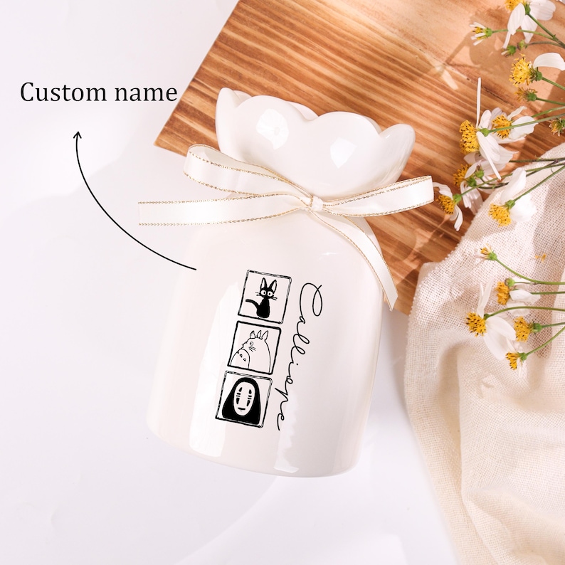 May include: A white ceramic vase with a scalloped top and a cream-colored ribbon. The vase features three black and white cartoon character illustrations and the word "Calliope" in script. A wooden surface and flowers are in the background.