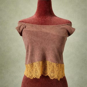 Puede incluir: Top marrón con hombros descubiertos y ribete de encaje amarillo mostaza. El top se exhibe en un maniquí rojo. El top tiene mangas cortas y un diseño sencillo, adecuado para ropa informal. El encaje añade un toque de elegancia.