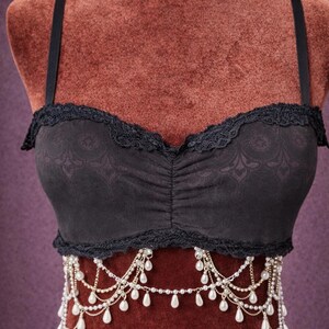 Op de afbeelding: Zwarte bustier met kanten rand en verstelbare bandjes. De bustier heeft een gerimpelde voorkant en is versierd met een delicate parel- en kettingversiering. Het kledingstuk wordt gepresenteerd op een mannequin met een bordeauxrode achtergrond.