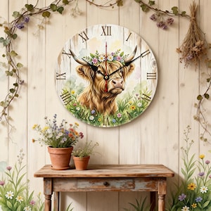 Op de afbeelding: Een ronde wandklok met een aquarel illustratie van een Schotse Hooglander versierd met een bloemenkrans. De klok heeft Romeinse cijfers en zwarte wijzers. De achtergrond is licht houtnerf.