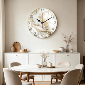 Puede incluir: Reloj de pared redondo con esfera de mármol blanco y vetas doradas. Los números negros marcan las horas, con manecillas negras y un segundero rojo. El reloj está montado en una pared de color neutro, sobre un mueble blanco y una mesa de comedor.