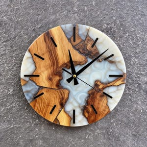Puede incluir: Reloj de pared redondo con un diseño único. La esfera del reloj presenta una combinación de madera marrón claro y resina blanca y azul. Las manecillas y los marcadores de hora negros completan el diseño. Un artículo de decoración.