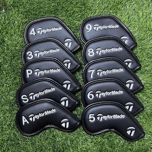 以下が含まれることがあります： 白の刺繍ロゴと文字が入った黒いTaylorMadeゴルフヘッドカバーのセット。各カバーには、4、9、3、8、P、7、S、A、6、5などの数字または文字が記載されています。緑の芝生の上に配置されています。