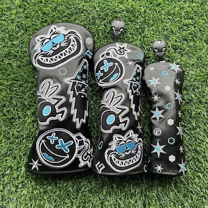 Puede incluir: Tres fundas negras para palos de golf con diseños de dibujos animados. Las fundas presentan un gato sonriente, una bomba con una carita sonriente y estrellas, todo en blanco y verde azulado. Las fundas están sobre un fondo de hierba verde.