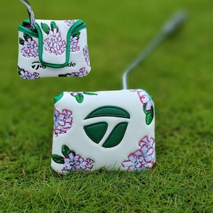 Puede incluir: Funda blanca para putter de golf con un estampado floral en rosa y morado, acentuado con ribetes verdes. La funda presenta el logotipo de TaylorMade en verde. La funda del putter se muestra sobre un fondo de hierba verde.