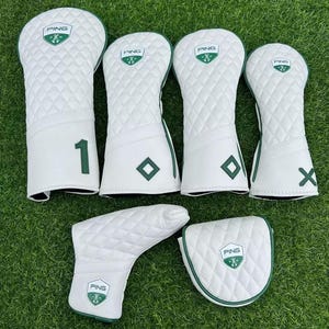 Puede incluir: Un conjunto de fundas blancas para palos de golf con un patrón acolchado y detalles en verde. Las fundas presentan el logotipo "PING" y varias marcas, incluyendo el número "1" y la letra "X". El conjunto incluye fundas para diferentes tipos de palos.