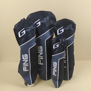 Puede incluir: Un conjunto de fundas para palos de golf en negro y gris. Las fundas presentan el nombre de la marca "PING" en blanco, junto con "G425". Están hechas de un material similar al cuero y tienen un diseño moderno.