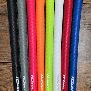 May include: A collection of eight golf club grips in various colors, including black, gray, pink, orange, green, white, red, and blue. Each grip has the word "IOMIC" printed in white. The grips are arranged on a wooden surface.
