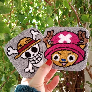 May include: Two gray, square, handmade coasters. One features a skull with a pirate hat and crossbones. The other depicts a cartoon character with a pink hat and antlers. The coasters are held in front of a green leafy background.