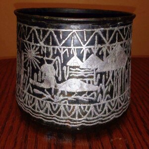 May include: A black metal cup with a silver design of a village scene. The design includes houses, trees, and a sun.