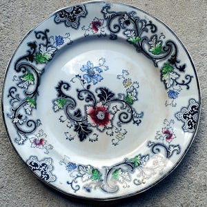May include: A white ceramic plate with a detailed floral design. The plate features a central floral arrangement with red, blue, and yellow flowers, surrounded by black and white scrollwork and green leaves. The rim is outlined in black.