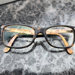 May include: Rectangular eyeglasses with a dark brown frame and patterned arms. The lenses are clear. The arms feature a light brown and beige design. The glasses are on a textured surface.