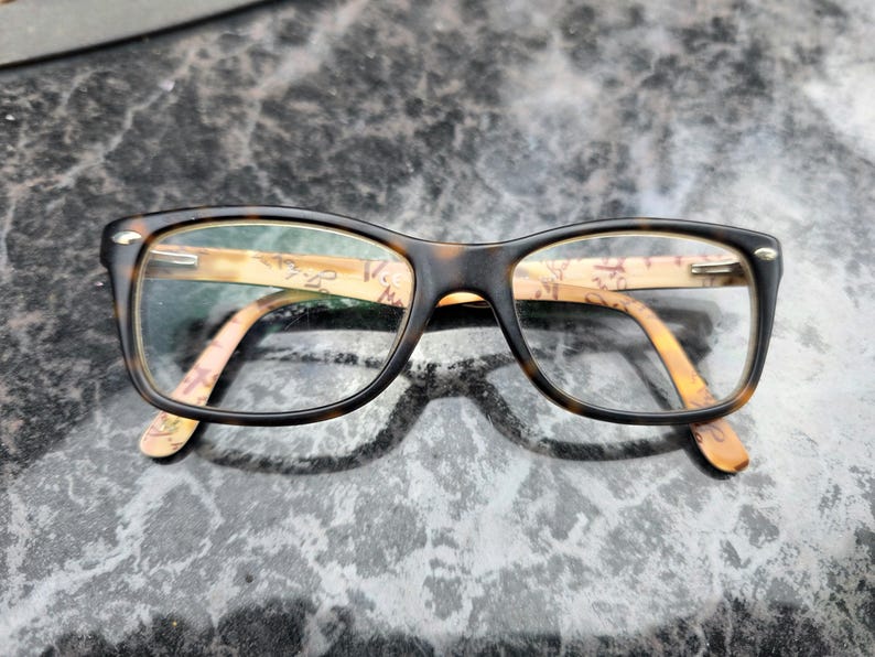 May include: Rectangular eyeglasses with a dark brown frame and patterned arms. The lenses are clear. The arms feature a light brown and beige design. The glasses are on a textured surface.