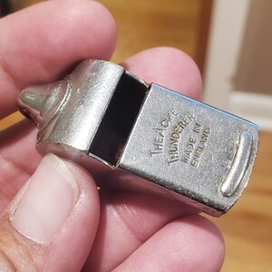 May include: A silver-colored Acme Thunderer whistle, made in England. The whistle has a rectangular body with rounded edges and a small mouthpiece. The text "The Acme Thunderer Made in England" is visible on the side.