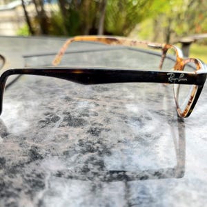 May include: Ray-Ban eyeglasses with a tortoiseshell pattern. The frames are dark brown on the arms, with the Ray-Ban logo visible. The lenses are clear. The glasses are designed for vision correction.
