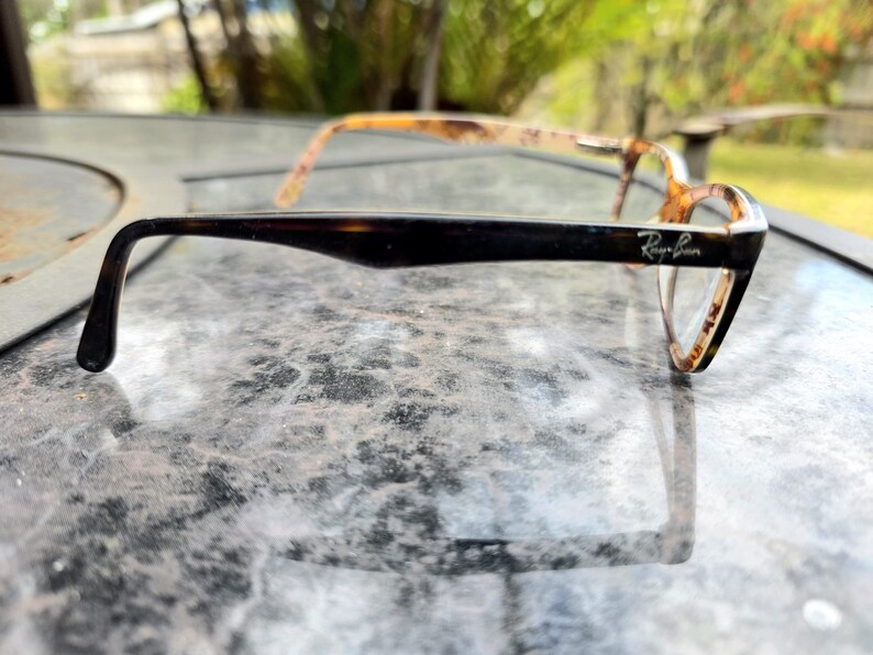 May include: Ray-Ban eyeglasses with a tortoiseshell pattern. The frames are dark brown on the arms, with the Ray-Ban logo visible. The lenses are clear. The glasses are designed for vision correction.