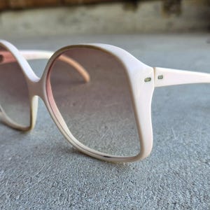 May include: Oversized vintage sunglasses with a light pink frame and gradient lenses. The sunglasses have a retro, square shape and are resting on a gray surface. The lenses transition from a darker shade at the top to a lighter shade at the bottom.