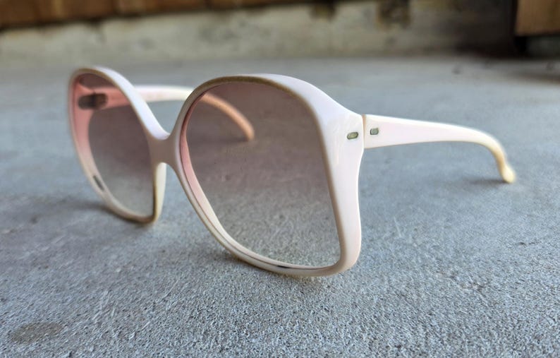 May include: Oversized vintage sunglasses with a light pink frame and gradient lenses. The sunglasses have a retro, square shape and are resting on a gray surface. The lenses transition from a darker shade at the top to a lighter shade at the bottom.