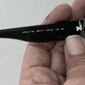 Op de afbeelding: Zwarte zonnebril met de tekst "MOD. 4136 GB1/11 64015 120 2N" in het wit gedrukt op de poot. De zonnebril wordt door een persoon vastgehouden.