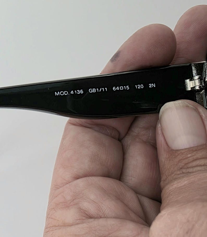 Op de afbeelding: Zwarte zonnebril met de tekst "MOD. 4136 GB1/11 64015 120 2N" in het wit gedrukt op de poot. De zonnebril wordt door een persoon vastgehouden.