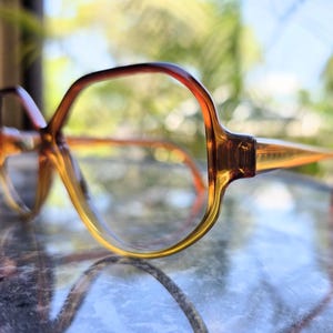 May include: A pair of vintage eyeglasses with a gradient amber and brown frame. The lenses are clear, and the glasses are resting on a reflective surface. The frames have a unique, angular shape, and the temples are a lighter amber color.