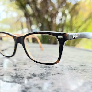 May include: Ray-Ban eyeglasses with a tortoiseshell frame. The rectangular glasses have clear lenses and a silver logo on the temple. The background is blurred, suggesting an outdoor setting. The frame is brown and black.