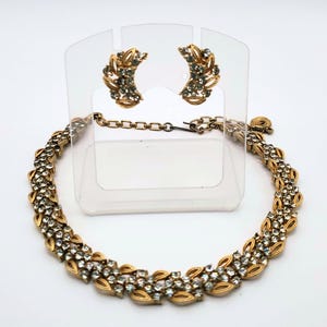 May include: A vintage jewelry set featuring a gold-toned necklace and matching clip-on earrings. The necklace and earrings are adorned with clear, sparkling rhinestones. The earrings are displayed on a clear stand. The necklace has a delicate chain and a hook clasp.