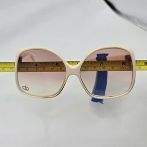 May include: Cream-colored oversized sunglasses with pink gradient lenses. The sunglasses are measured with a yellow tape measure. The frame has a small emblem on the left lens. The sunglasses are approximately 6 inches wide.