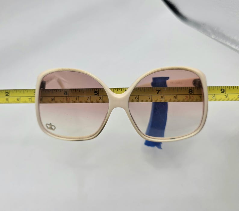 May include: Cream-colored oversized sunglasses with pink gradient lenses. The sunglasses are measured with a yellow tape measure. The frame has a small emblem on the left lens. The sunglasses are approximately 6 inches wide.
