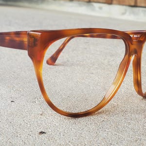 May include: A pair of vintage eyeglasses with a tortoiseshell pattern. The frames are a warm brown color with a rounded, oversized shape. The temples and bridge are a darker shade of brown.