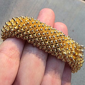 Puede incluir: Un brazalete dorado con una superficie texturizada compuesta por numerosas cuentas esféricas pequeñas. El brazalete se sostiene en una mano, mostrando su intrincado diseño y brillo metálico. La pieza de joyería parece ser un accesorio llamativo.